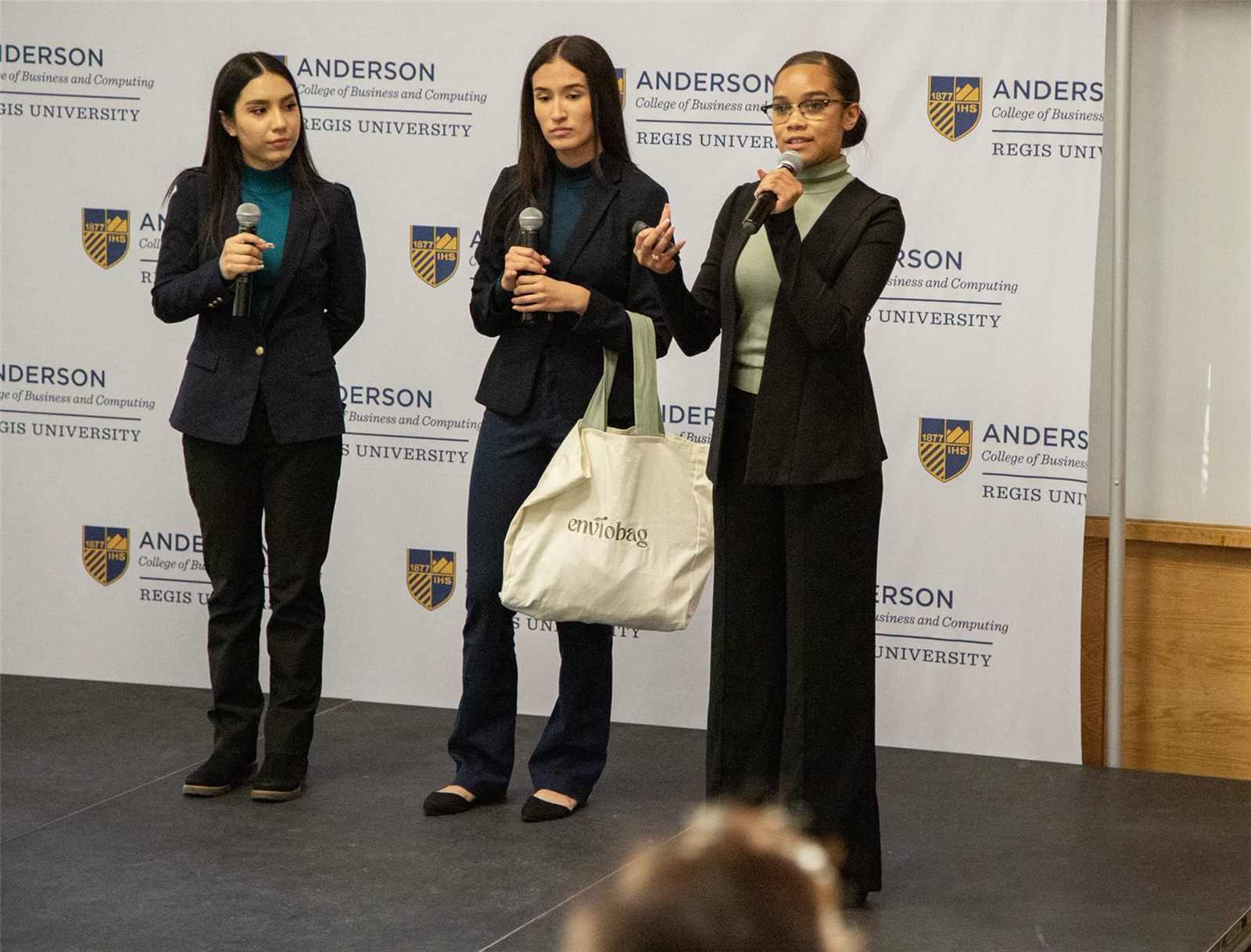 Three young women in blazers pitch on stage. One holds a canvas tote printed with “enviobag.”