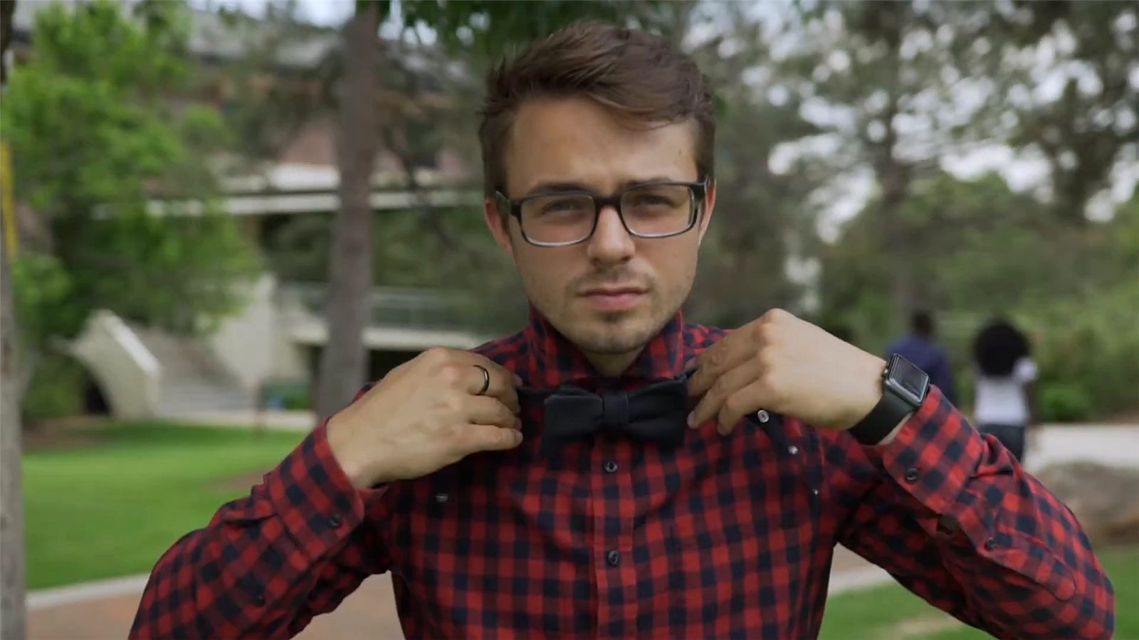 Paul Hunter, in glasses and a red plaid shirt, straightens his dark bowtie on the Regis campus