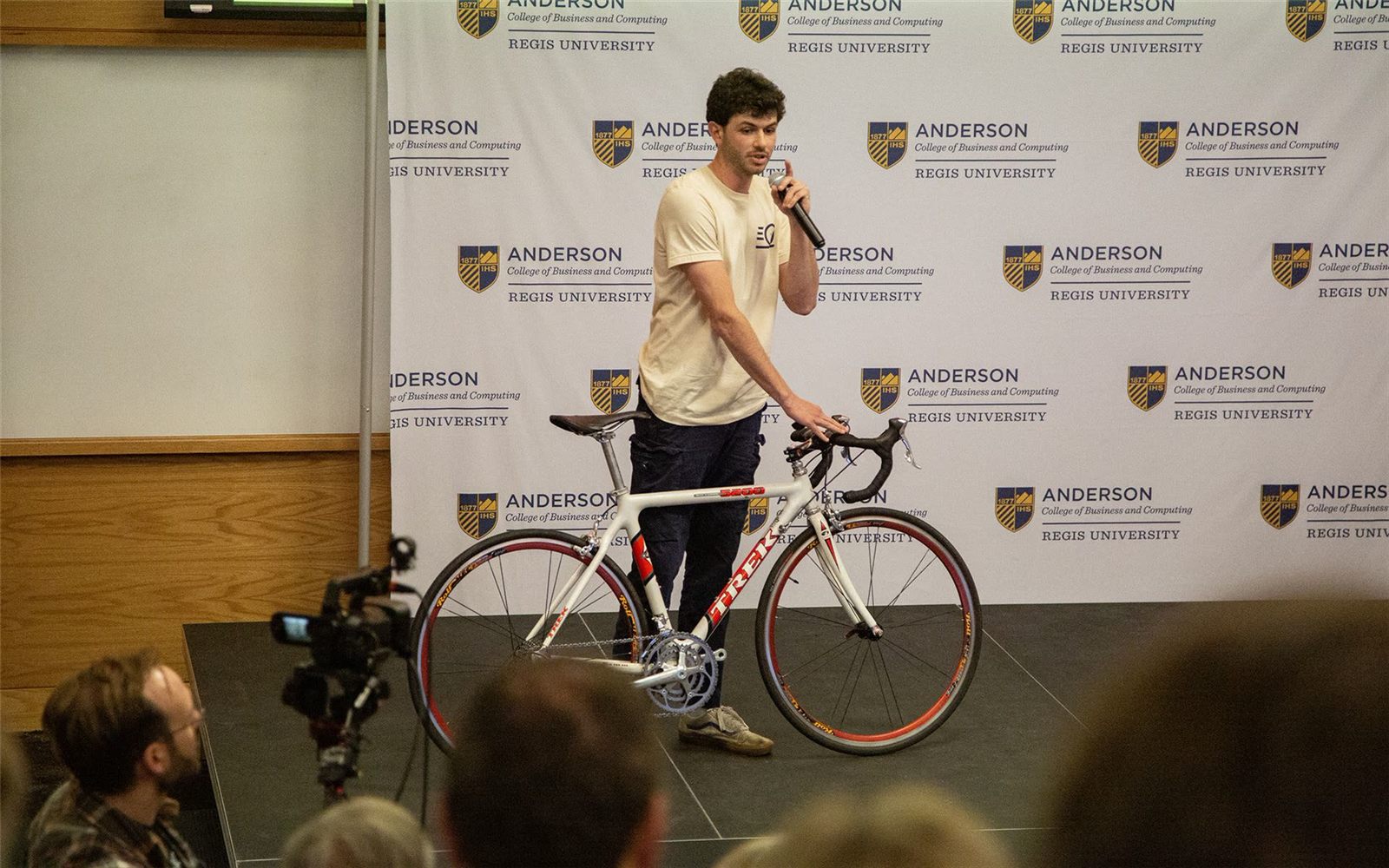 Anton Cariffe, in a Traveling Ground T-shirt, pitches on the Anderson College stage with a hand on a white Trek road bike.